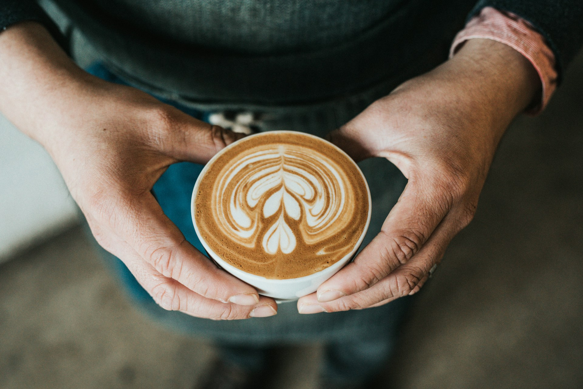 A person holding a cup of coffee in both hands, viewed from above, with a foam design of three hearts stacked vertically