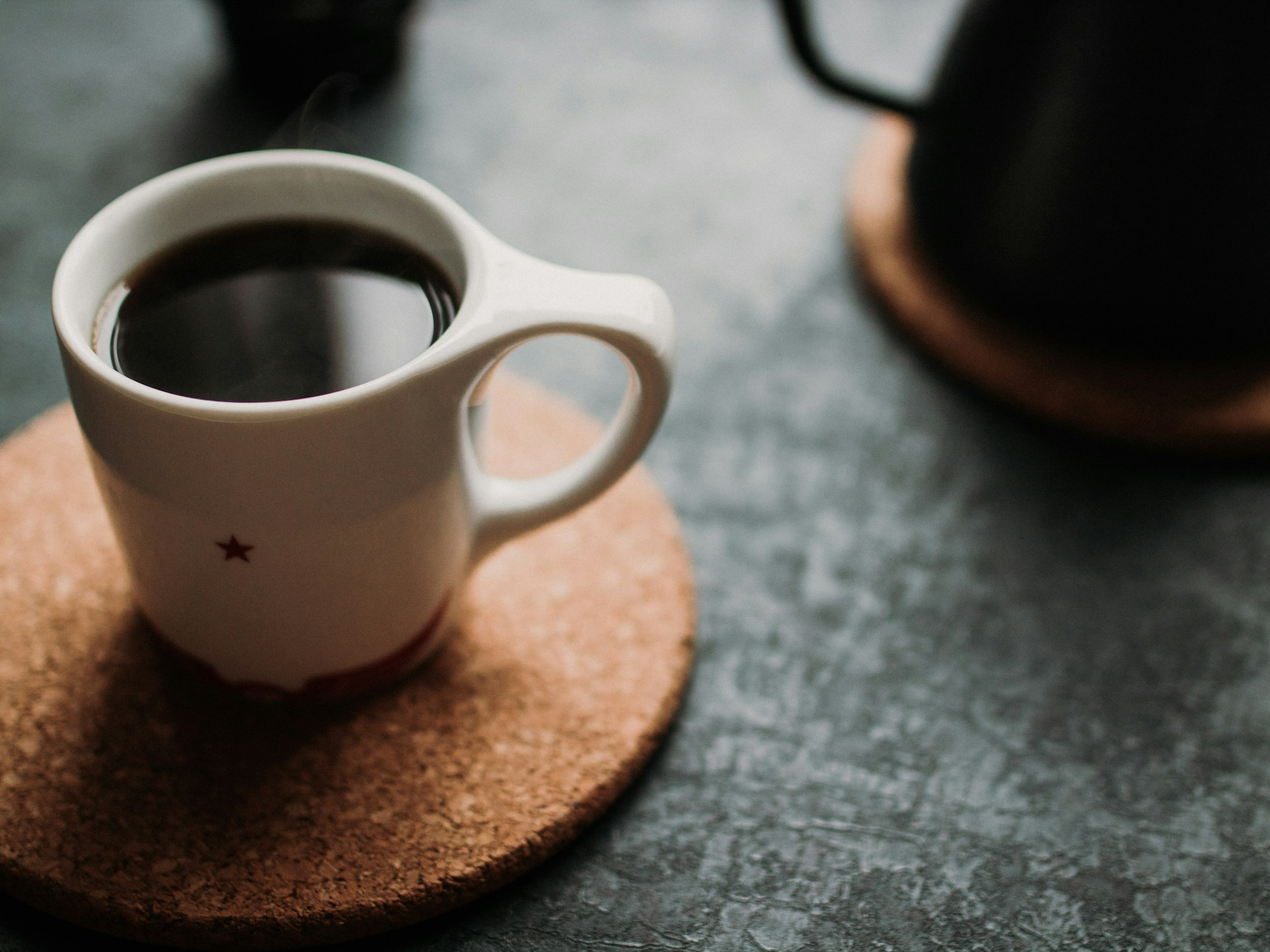 A cup of black coffee in a white mug with a little star on it, sitting on a large cork mat