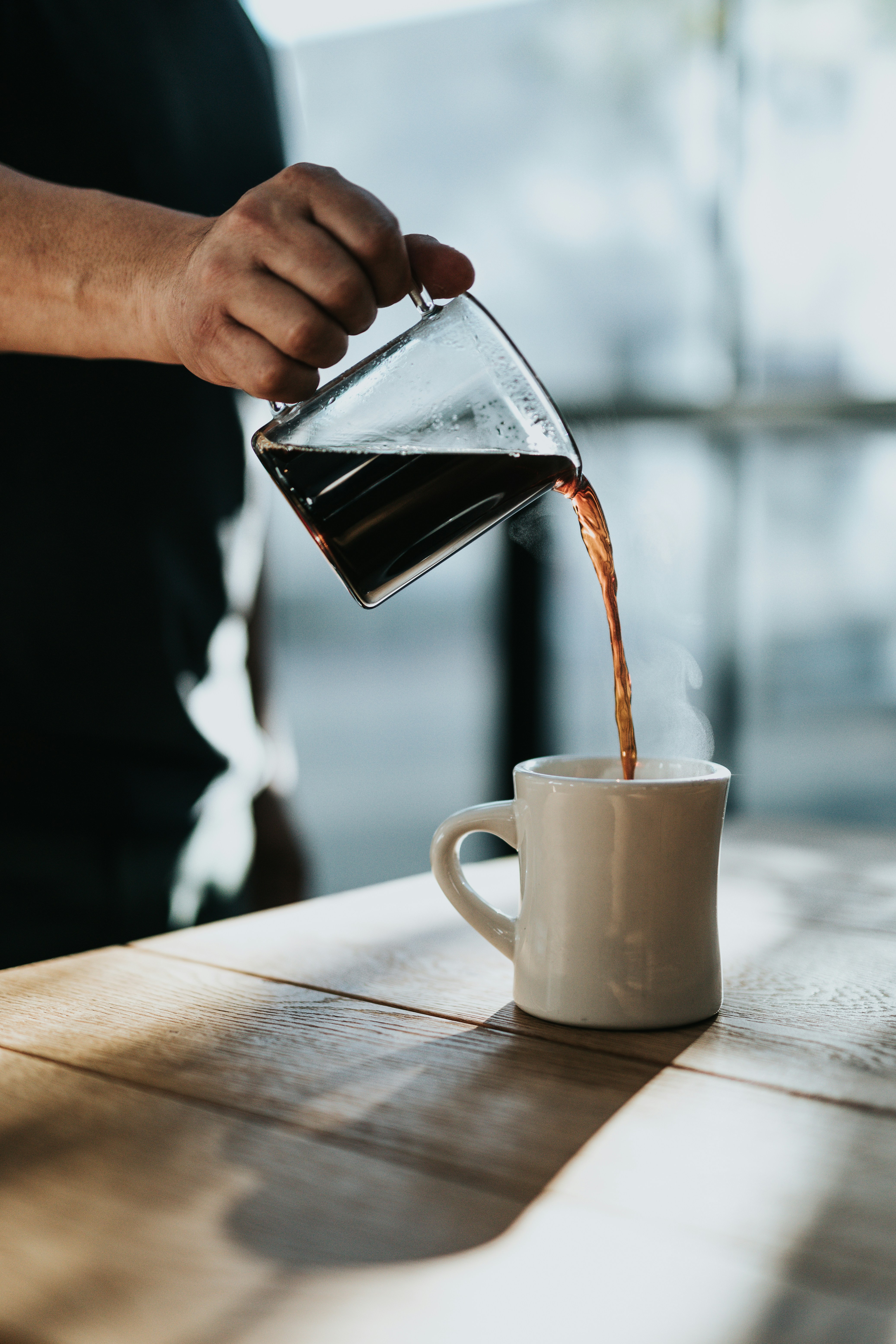 A person wearing all black, pouring black coffee into a white mug