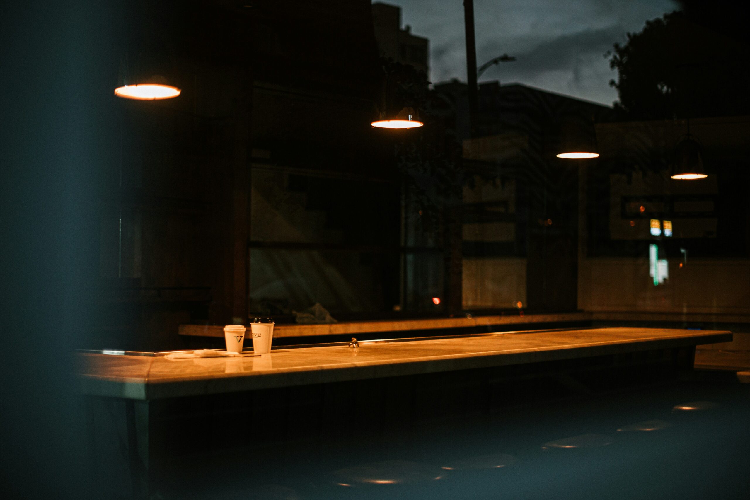 A pair of to-go mugs sit on a table illuminated by orange overhead lamps while the sky outside is dark